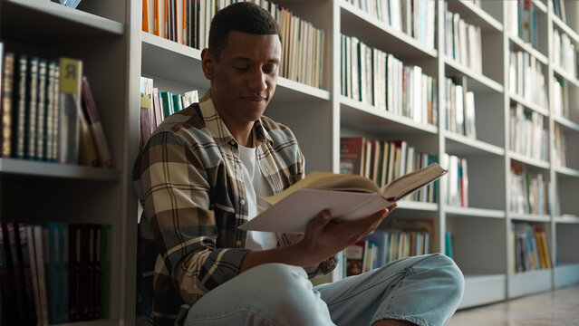 African American man sitting library floor bookcase reading book pleasure hobbies story knowledge learning student turning pages male guy academy study literary university history interesting reader