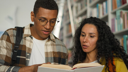 Two students African American man Arabian Latino woman together reading book textbook library talking discussion university college classmates speak study learning hobby girl guy high school bookstore