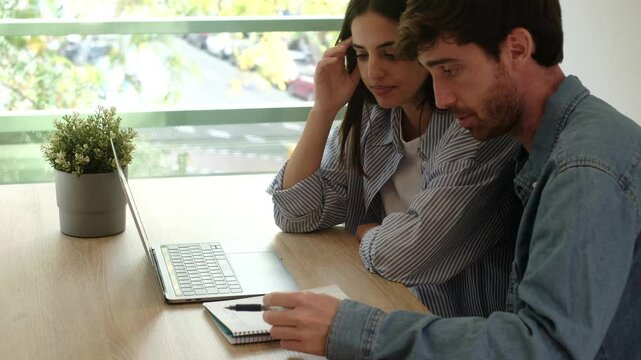 Young couple discussing their household finances, sitting at the kitchen table with a laptop, reviewing monthly bills, utility payments, and calculating how to balance their limited income with daily 