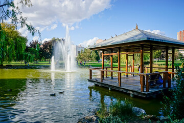 Park with a lake that has a vertical water jet and a wooden pergola