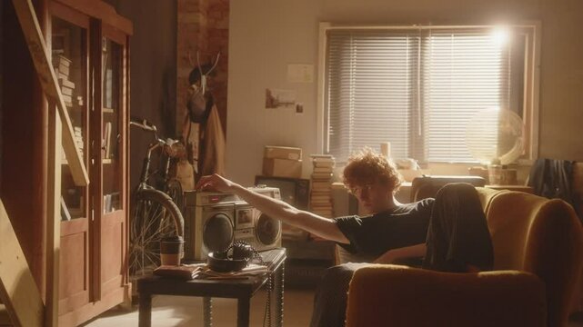 Melancholic young man lounging on sofa in warmly lit room, inserting audio cassette and adjusting volume on vintage boombox, listening to music to solace from his thoughts at home. Zoom shot
