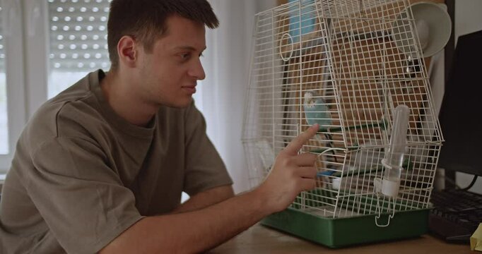 A young man interacts with his pet parakeet in its cage at home. He gently strokes the bird, showing affection and care.