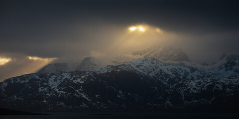 A breathtaking view of Lofoten, Norway, during golden hour. Snow-covered peaks surround a still fjord, with sun rays piercing through the clouds, illuminating the Arctic winter landscape.
