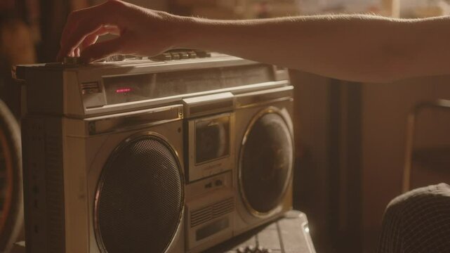 Young sad man sitting on couch in sunlit room, inserting audio cassette and turning on music on retro boombox, trying to escape from his thoughts. Close-up shot