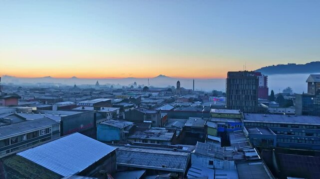 Toma a&eacute;rea de amanecer en Temuco con volcanes de fondo y edificios peque&ntilde;os cubiertos de niebla matutina grabado con dron DJI mavic 3 resoluci&oacute;n ultra hd Temuco sur de Chile 