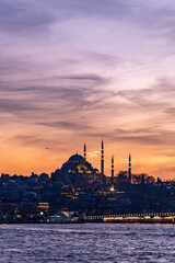 High resolution panoramic photograph capturing Istanbul's historic peninsula bathed in a crimson sunset glow. The city's iconic skyline silhouettes against the vibrant evening sky