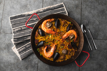Classic Spanish dishes, seafood paella in a traditional skillet against the rustic blue concrete backdrop. Spanish paella with shrimp, hugs, mussels, green peas, and fresh lemon wedges from above.