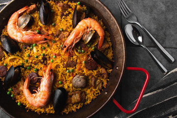 Classic Spanish dishes, seafood paella in a traditional skillet against the rustic blue concrete backdrop. Spanish paella with shrimp, hugs, mussels, green peas, and fresh lemon wedges from above.