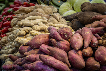A full frame photograph of a selection of fresh produce for sale on a market stall, with sweet potatoes in the foreground