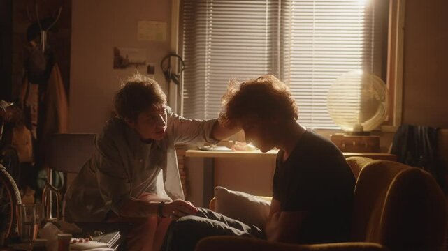 Sad young man sitting hunched on sofa in warmly lit living room, his concerned girlfriend comforting and supporting him during difficult times or depression. Zoom shot