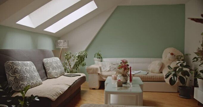 Young Woman Relaxes on Sofa, Browsing Smart phone in Cozy Attic Apartment. Natural light from skylights illuminates the scene, creating a warm, inviting atmosphere.