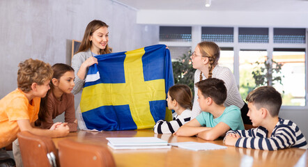 Decent teacher showing Sweden flag to group of preteen schoolchildren in classroom during lesson