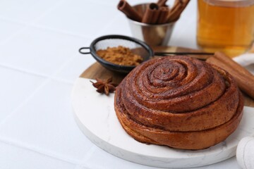 Delicious cinnamon roll bun, tea and spices on white tiled table, closeup