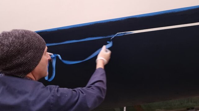 A man working on a boat in a boatyard peeling off masking tape from the hull after painting revealing a clean white line finish.