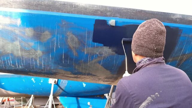 A man working in a boatyard painting antifoul paint onto a sailing yacht boat with a small roller.