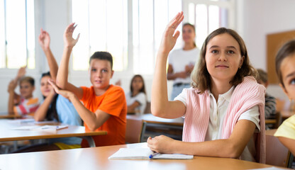 Portrait of diligent interested tween schoolgirl sitting with hand raised during lesson in classroom