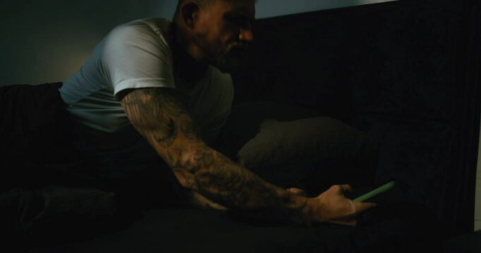 A guy with tattoos in a white T-shirt looks at the alarm clock on his smartphone and wakes up in bed in the morning