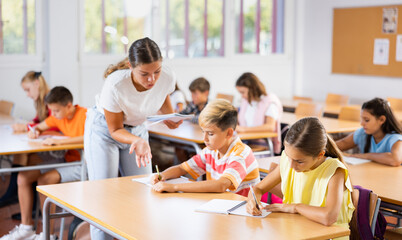 Positive boys and girls studying and woman teacher helping in classroom