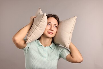 Annoyed woman covering her ears with pillows due to loud sound on light grey background