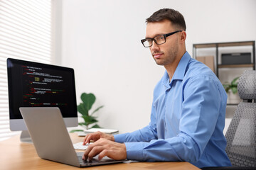 Programmer working on laptop and computer at wooden desk indoors