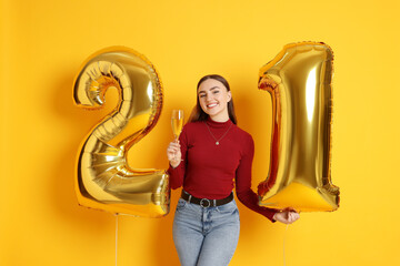 Coming of age party - 21st birthday. Happy young woman with number shaped balloons and glass of sparkling wine on yellow background