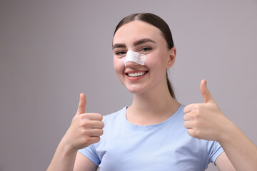 Woman with medical bandage on her nose after plastic surgery operation showing thumbs up against...