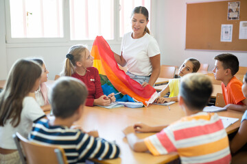 Schoolchildren sitting in classroom and listening to female teacher. She holding rainbow flag in hands and talking about minorities