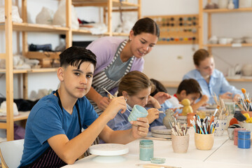 Teenager paints a clay mug with brush at a workshop with a teacher. Boy at a ceramics master class works at a table in an art studio
