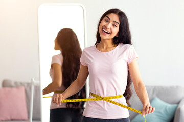 Millennial Indian woman measuring her waist with tape in front of mirror, showing results of...