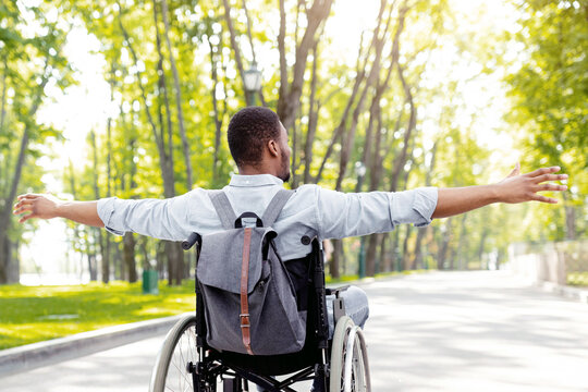 Back view of young disabled black man in wheelchair spreading his arms, feeling happy and free on walk at city park. Full length of handicapped guy with backpack going on trip outdoors