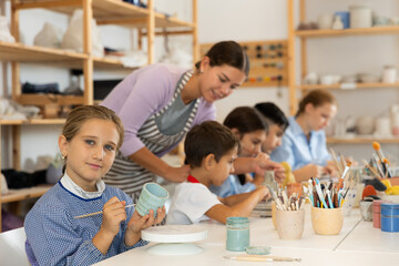 Focused girl paints a ceramic glass made for a master class with a brush. Children make pottery at a workshop in a creative studio