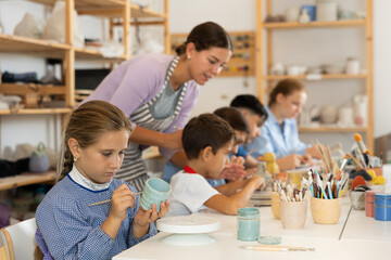 Interested little girl painting handmade ceramic cup with brush in pottery workshop