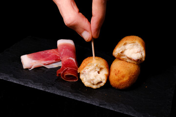 Hand picking a ham croquette with slices of cured ham on a slate plate