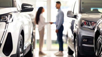 Unrecognizable young middle eastern man and woman shaking hands at newest auto showroom, blurred background. Customer buying brand new luxury car at automobile dealership salon