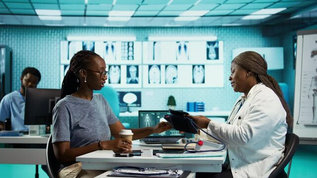 Black medic practitioner takes blood pressure with a blood pressure monitor, does a consultation with female patient using modern advanced tools for the checkup routine. Camera B.