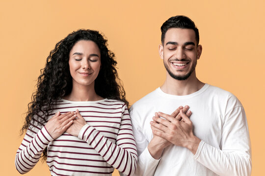 Thank You. Grateful Young Arab Man And Woman With Eyes Closed Keeping Both Hands On Chest, Thankful Middle Eastern Couple Expressing Grattitude And Kindness While Posing Over Yellow Background
