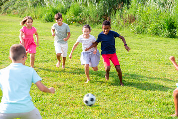 Obraz premium Happy preteen girls and boys of different nationalities playing football on green lawn in spring city park..