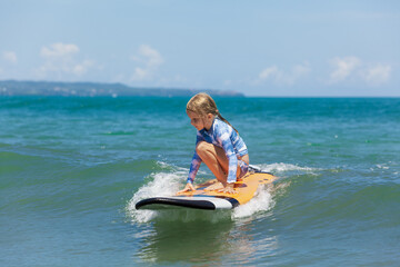 Happy baby girl - young surfer ride on surfboard with fun on sea waves. Active family lifestyle, kids outdoor water sport lessons and swimming activity in surf camp. Summer vacation with child.