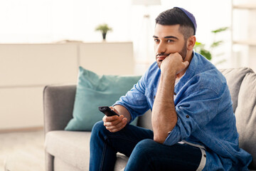 Fototapeta premium Boring Television Program. Bored Jewish Man Watching TV Switching Channels With Remote Control Sitting On Sofa At Home. Discontented Viewer Watching Uninteresting Movie, Feeling Lonely