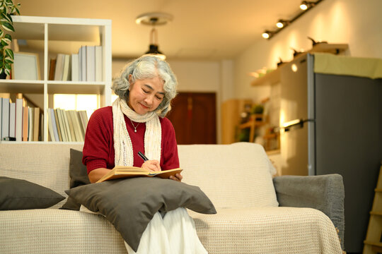 Calm senior woman in a red sweater sitting on couch and writing in journal