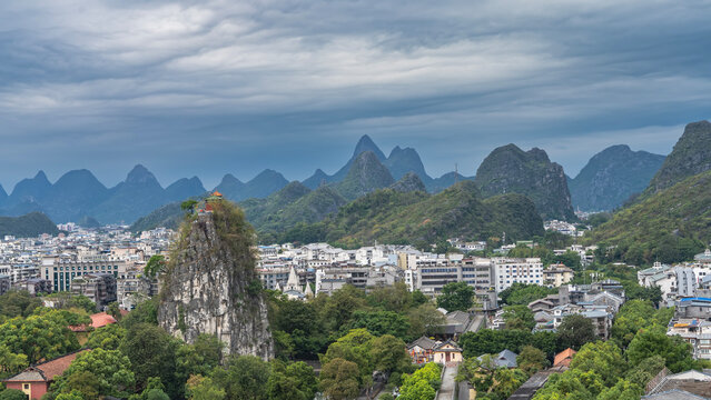 Beautiful urban and mountain scenery. City buildings surrounded by green trees. A high cliff with steep slopes and a pavilion at the top. A mountain range against the sky and clouds. China. Guilin.