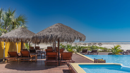 Fototapeta premium Sun beds under straw umbrellas on the boardwalk next to the swimming pool. There is a sandy beach and green vegetation ahead. The ocean is far away. Clear blue sky. Copy space. Madagascar. Morondava.