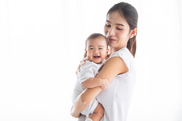 Asian beautiful young single mother holding smiling newborn baby in bright indoor. Both dressed in white, sharing joyful and affectionate moment home environment. Baby healthcare products concept