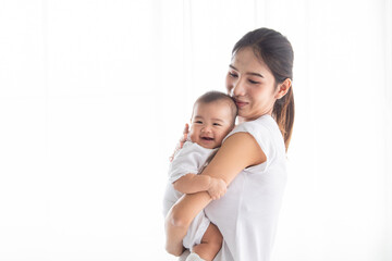 Asian beautiful young single mother holding smiling newborn baby in bright indoor. Both dressed in white, sharing joyful and affectionate moment home environment. Baby healthcare products concept
