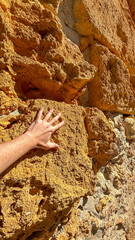 A close-up of a hand gently resting on a weathered stone wall, highlighting the texture and rich orange hues of the surface. The sun casts a warm glow on the stone.