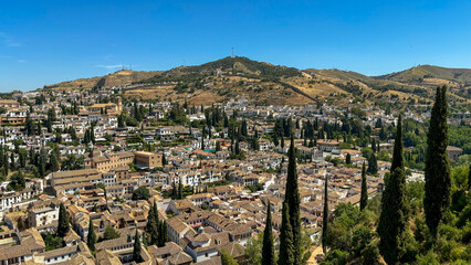 A scenic view of the Albayzín district in Granada, Spain, showcasing its traditional whitewashed houses, cypress trees, and rolling hills under a clear blue sky.