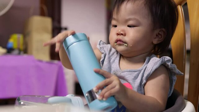 Baby girl enjoys her mealtime, holding a food container and sippy cup while sitting on a high chair