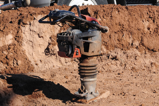 An idle tamping rammer vibratory asphalt, soil plate compactor on a construction site, positioned in a dirt area with visible layers of excavated earth in the background