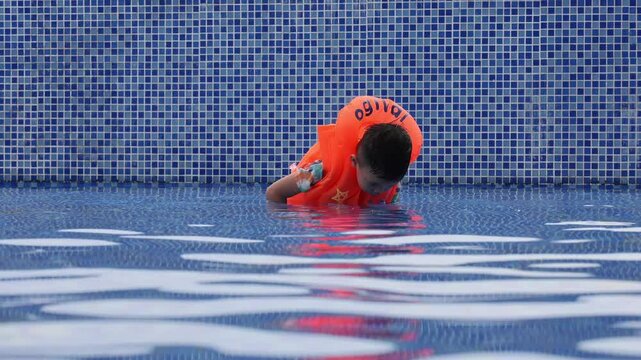 Young boy wearing orange floaties practices holding his breath underwater in a pool with blue tile