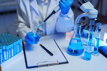 Scientist in a lab coat and gloves pouring blue liquid from a micropipette into a beaker,...